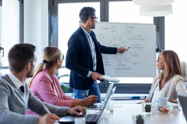 Formateur en réunion, présentant une stratégie sur tableau blanc à des dirigeants en formation, session de formation soft skills dirigeants en entreprise.